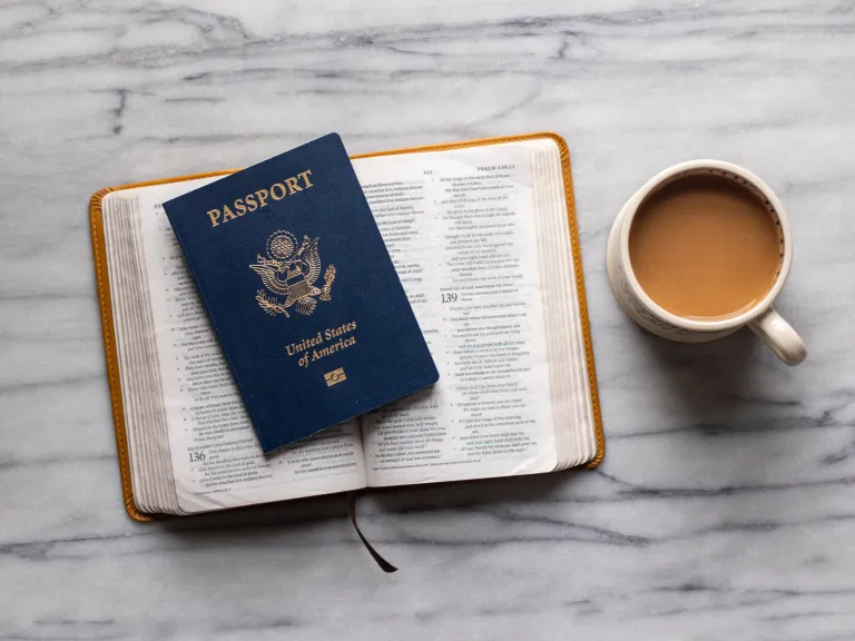Our passports are physical proof of who we are and to what country we belong. a open Bible with a US passport placed on top sitting beside a mug of coffee on a marble tabletop